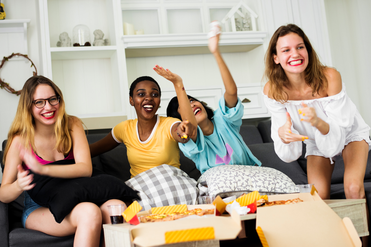 A group of diverse woman friends sitting on the couch eating pizza together.