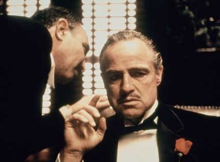 Vito Corleone is seated on one corner and an unknown person comes to whisper something to his ear.